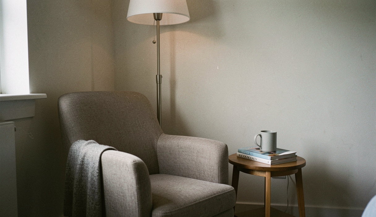 A low armchair in the corner of a room with a floor lamp behind it and a small side table holding a mug and a paperback