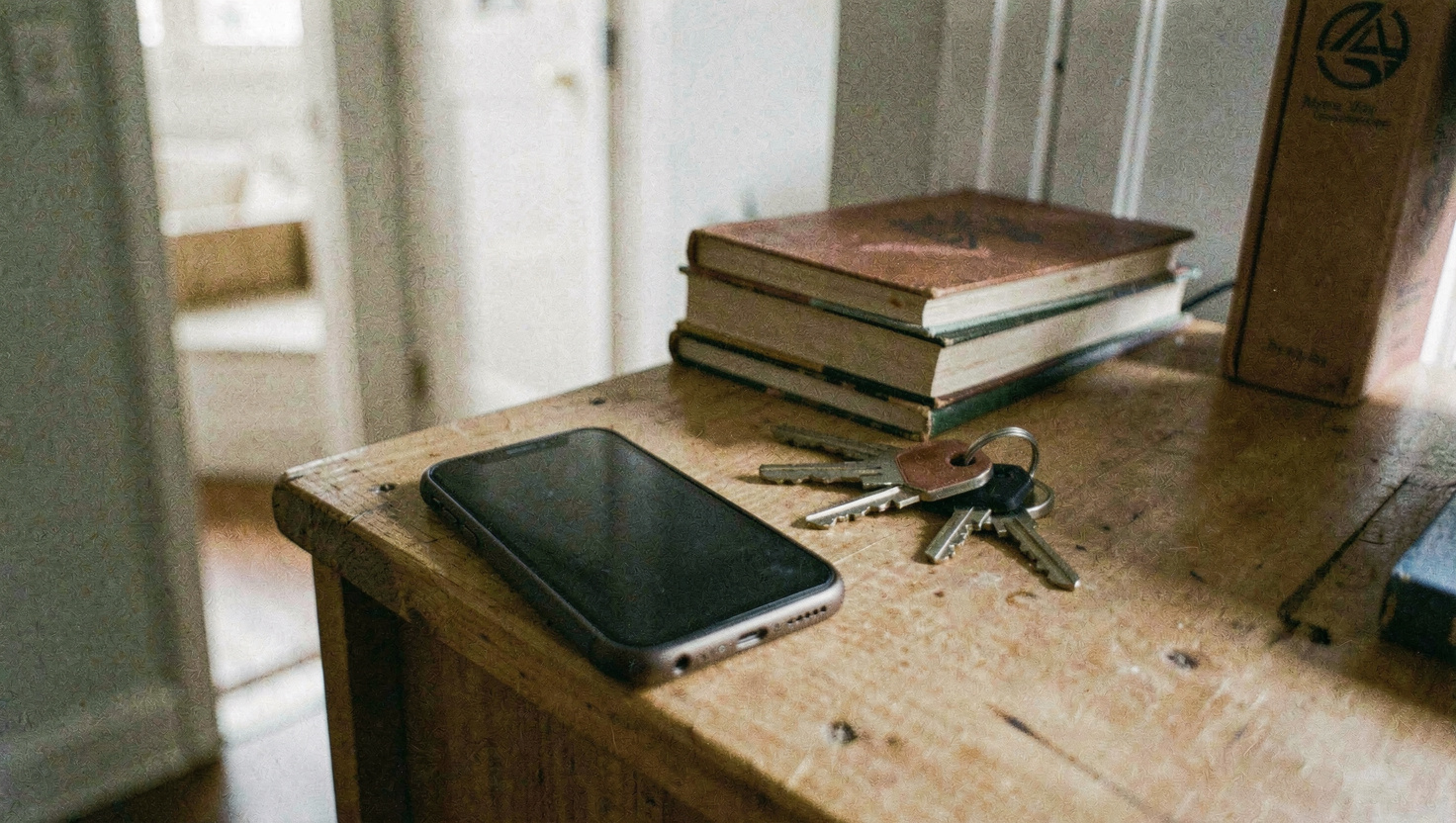 A phone lying on a wooden shelf beside a small stack of books and a pair of keys, the screen dark, a doorway visible beyond